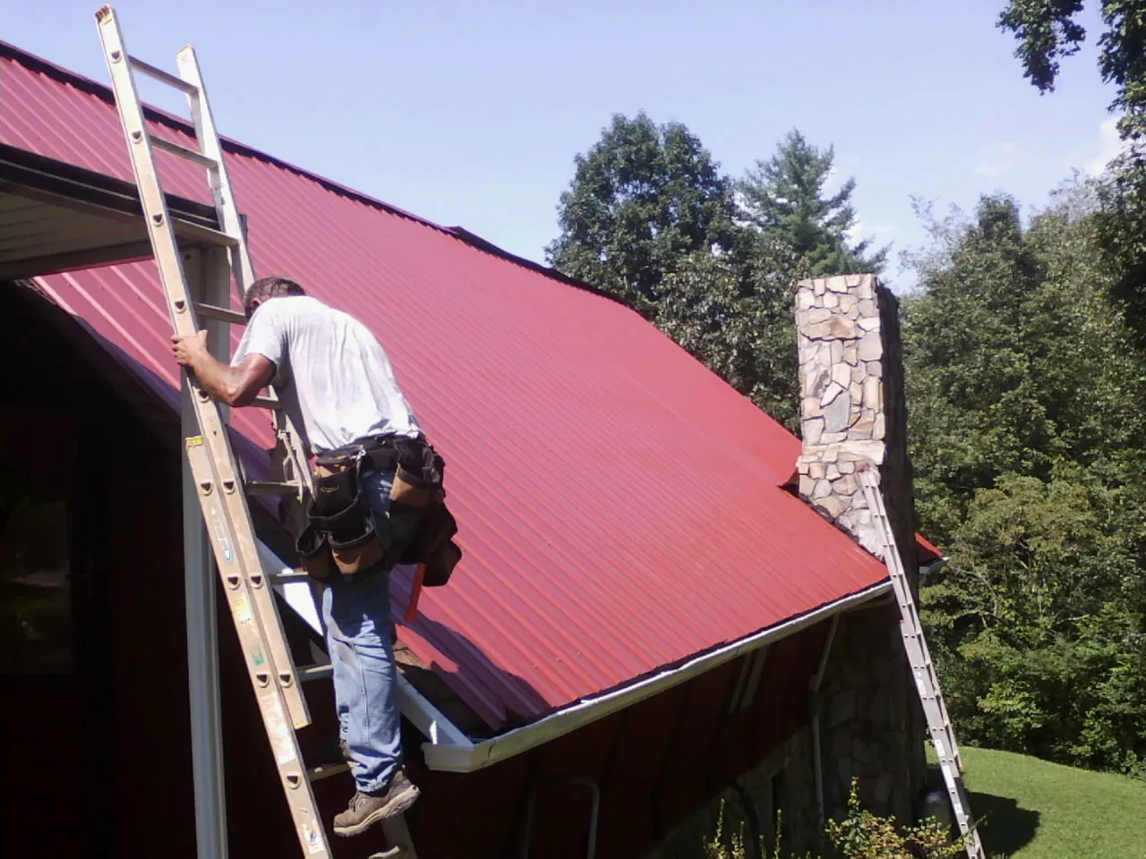 Roofer on a ladder beside a red metal roof and stone chimney — Soffit & Fascia Repair by Roofing Brothers in Treadwell